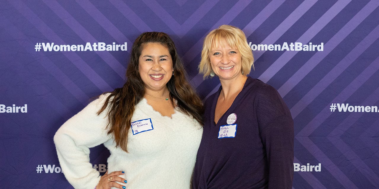 Two Silver Lakeassociates in front of a purple banner with #womenatbaird hashtags