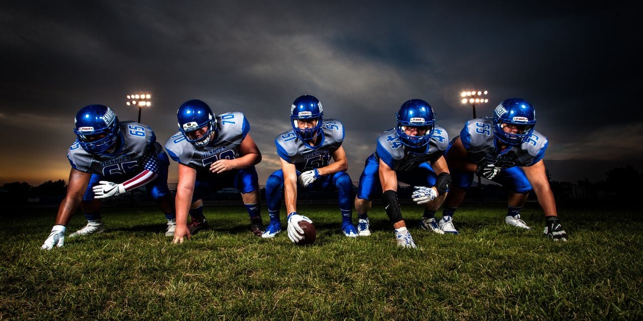 Five football players in blue uniforms lined up on a field at dusk, ready for a play.